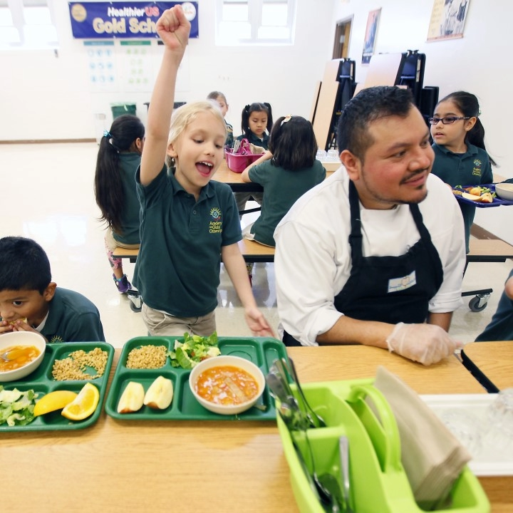 A Cultivate Collective chef sits with children in the lunchroom, the child next to him is excitedly jumping up with an arm in the air